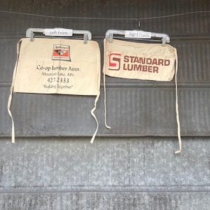 May include: Two tan aprons hanging on a metal rod. The left apron has the text "Co-op Lumber Assn." and the right apron says "STANDARD LUMBER". Both aprons have a pocket and tie strings.
