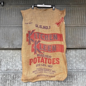 May include: A burlap potato sack with red text that reads "Kitchen Kleen Washed Potatoes." The sack is labeled "U.S. No. 1" and states the net weight is 100 lbs. The sack is hanging from a black hanger.