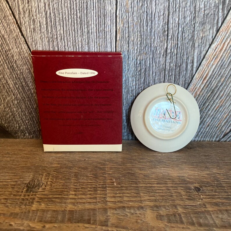 May include: A white porcelain plate with a red and black image of 101 Dalmatians. The plate has a gold hook on the back and is dated 1996.