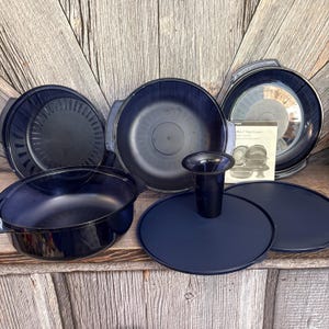 May include: A dark blue Tupperware Microwave Stack Cooker set including multiple round containers, lids, a cone-shaped insert, and an instructional booklet. The components feature smooth, deep blue plastic surfaces and integrated handles for easy stacking.
