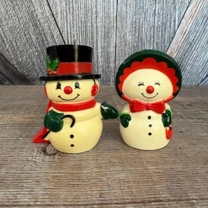 May include: Two ceramic snowman figurines. One snowman wears a black top hat with holly, a red scarf, and green mittens. The other snowman has a green hat with a red trim, a red bow tie, and green mittens. Both have black button eyes and smiles.