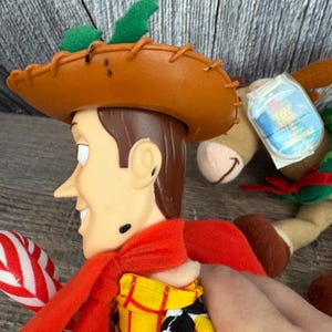May include: A Woody doll from the Toy Story franchise. The doll has a brown cowboy hat, a red scarf, and a yellow shirt with a black and white pattern. The doll is holding a red and white striped candy cane.