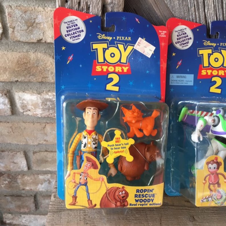 May include: Two Toy Story 2 action figure sets in their original packaging. One features Woody, a cat, and a bear. The other shows Buzz Lightyear and a monkey. The packaging is blue with the Toy Story 2 logo.