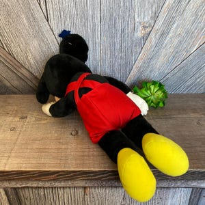 May include: A black plush Mickey Mouse doll wearing red dungarees is lying on a wooden surface. The doll has yellow shoes and a blue bow on its head.