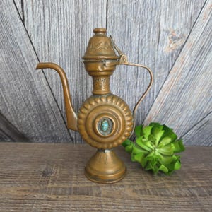 Puede incluir: Una tetera antigua de color cobre con un pico largo y un asa. La tetera tiene un cuerpo redondo con un diseño circular decorativo y una pequeña piedra turquesa en el centro. La tapa es abatible y tiene un patrón decorativo.