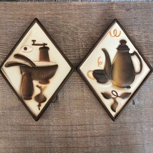 May include: Two ceramic wall tiles with a diamond shape. The first tile features a brown and white illustration of a mortar and pestle, a garlic bulb, and a grinder. The second tile features a brown and white illustration of a teapot, a cup, and a leaf.