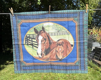 May include: A blue and green plaid fabric with a central oval image of a brown horse and foal. The horse has a white blaze. The background features a fence and trees. The fabric is hanging outside on a clothesline.