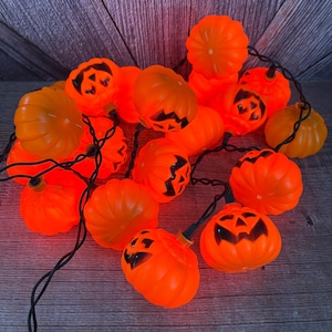 May include: A string of orange pumpkin-shaped lights connected by a black wire. The set features a mix of plain ribbed pumpkins and pumpkins with black jack-o'-lantern facial features, including triangular eyes and jagged mouths.