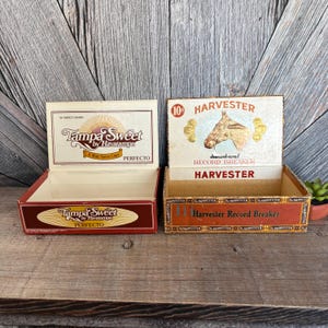 May include: Two vintage cigar boxes. The left box is labeled "Tampa Sweet by Havatampa" with "PERFECTO" text. The right box is labeled "HARVESTER" with a horse graphic and "Record Breaker" text. Both boxes are open, showing their interiors.