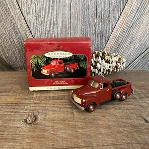 May include: Red 1953 GMC All-American Truck Hallmark Keepsake Ornament. The truck is carrying a small Christmas tree. The ornament is in its original box.