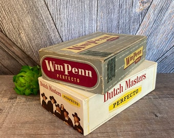 May include: Two vintage cigar boxes stacked on top of each other. The top box is gray with the text "Wm Penn Perfecto" in red and white. The bottom box is white with the text "Dutch Masters Perfecto" in black and yellow.