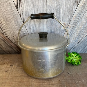 May include: A vintage aluminum camping pot with a black handle and lid. The pot has a black knob on the lid and measurement markings on the side.
