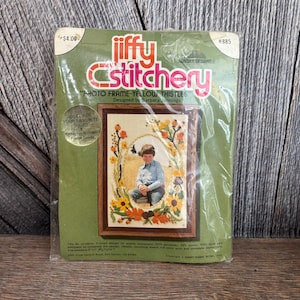 May include: A Jiffy Stitchery photo frame kit featuring a design of yellow thistles. The kit includes a printed design, needle, mounting board, and full-color print. The frame is designed by Barbara Jennings and is packaged in a green plastic sleeve.
