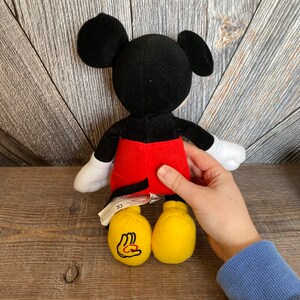 May include: A plush Mickey Mouse toy with black fur, red shorts, and yellow shoes. The toy is standing on a wooden surface.