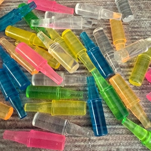 May include: A collection of colorful, transparent plastic bottle stoppers in various shades including pink, yellow, green, and blue. The stoppers are scattered across a textured surface, showcasing their varied hues and shapes.