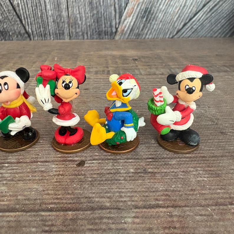 May include: Four miniature Christmas figurines featuring Mickey Mouse, Minnie Mouse, and Donald Duck. Mickey and Minnie are wearing red and white outfits with Santa hats. Donald Duck is wearing a blue jacket and Santa hat. Each figurine is on a penny.