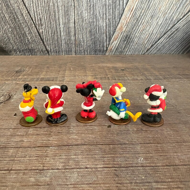 May include: A collection of five miniature Christmas-themed Disney character figurines. Mickey Mouse, Minnie Mouse, Donald Duck, and Pluto are dressed in festive holiday attire, with Santa hats and winter clothing. Each figurine is approximately 2 inches tall.