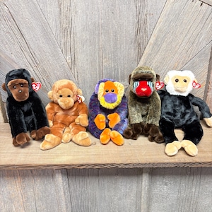 May include: Five plush monkey toys in various colors sit on a wooden shelf. The monkeys include black, brown, orange, purple, and green. Each monkey has a different facial design and a red heart-shaped tag.