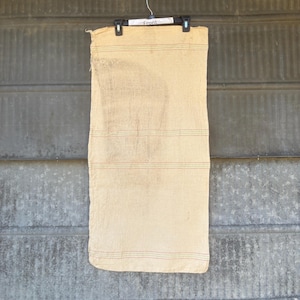 May include: A tan-colored, rectangular textile with horizontal red and green stitching. The textile is hanging from a black hanger with the word "Front" printed on it. The background is a corrugated metal surface.