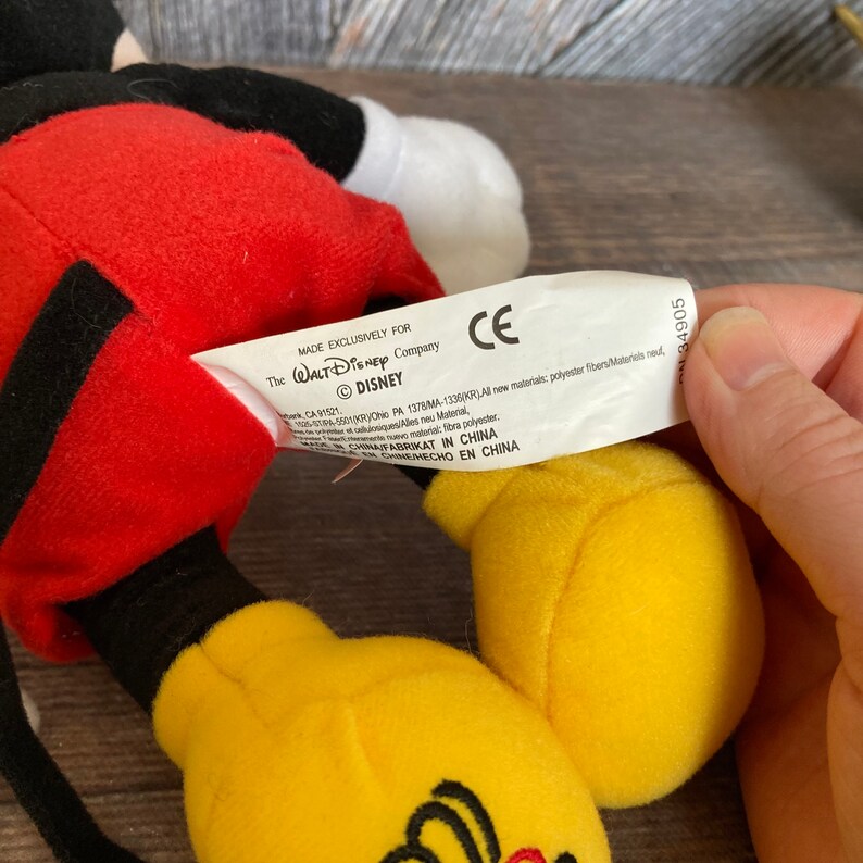 May include: A red and yellow plush Mickey Mouse toy with a black outline. The toy has a tag that reads "Made Exclusively for The Walt Disney Company &copy;DISNEY".