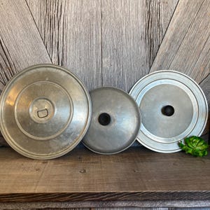 May include: Three silver metal lids with black knobs. The lids are different sizes and are arranged in a row on a wooden surface.
