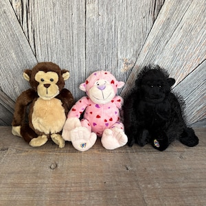 May include: Three plush monkey toys. The first is brown with a tan face and ears. The second is pink with heart patterns and the letter "W" on its foot. The third is a black gorilla.