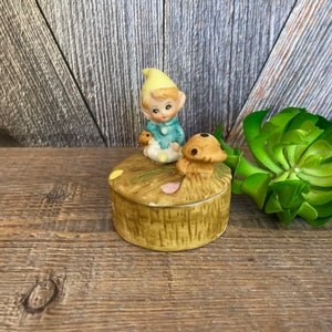May include: A ceramic trinket box featuring a small elf figurine sitting on a mushroom. The box is decorated with brown, green, and yellow accents.