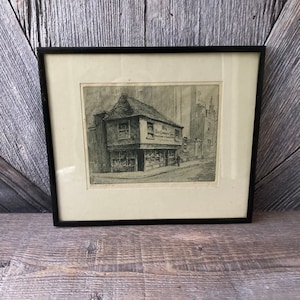 May include: Black framed print of a detailed street scene. The artwork depicts a two-story building with the text "The Old Curiosity Shop" on the side. A person walks on the street in front of the building. The print is on a white mat.