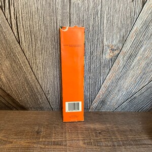 May include: An orange rectangular box with the text "CAUTION: NEVER LEAVE BURNING CANDLE UNATTENDED" printed on it. The box is on a wooden surface with a barcode at the bottom.