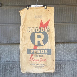 May include: Tan feed sack with the text "RODDIS" above a large blue "R" inside a circle, with a red lightning bolt design. Below is "FEEDS" and "100 LBS." The words "Always Fresh" are written in cursive. The bag is hanging from a black hanger.