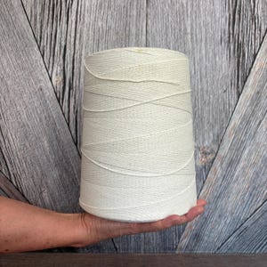 May include: A large spool of off-white twine, held in a person's hand. The twine is tightly wound, creating a conical shape. The background is a weathered, gray wooden surface.
