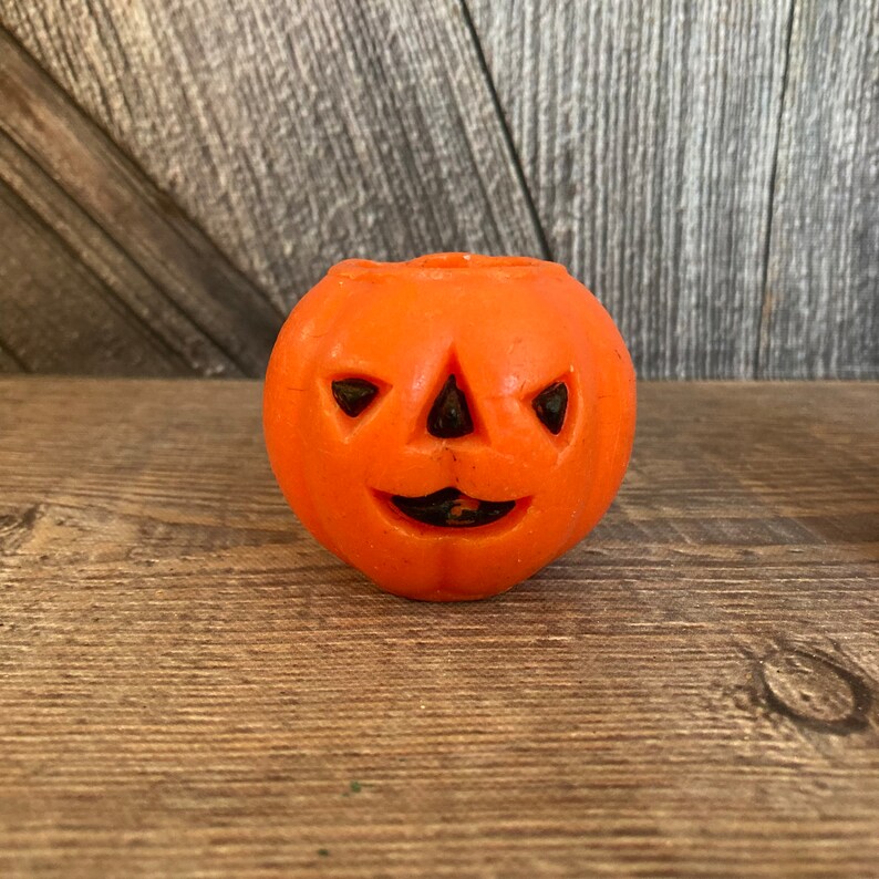 May include: Orange pumpkin-shaped candle with a carved jack-o'-lantern face. The candle has a black triangular nose and eyes.