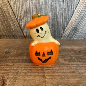 May include: A white ghost candle with a smiling face wearing an orange pumpkin hat. The ghost is sitting on top of an orange pumpkin candle with a carved jack-o'-lantern face.
