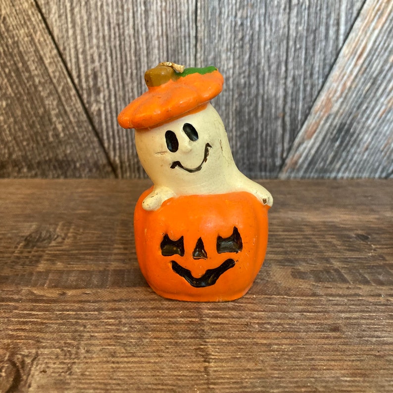 May include: A white ghost candle with a smiling face wearing an orange pumpkin hat. The ghost is sitting on top of an orange pumpkin candle with a carved jack-o'-lantern face.