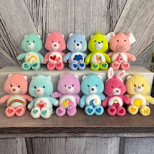 May include: A collection of eleven plush Care Bears in various colors, including pink, blue, yellow, and green. Each bear features unique belly badges, such as hearts, rainbows, and clouds. The bears are arranged on a white surface against a wooden backdrop.