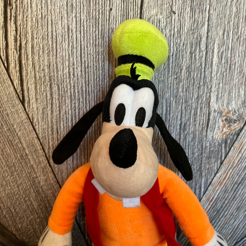 Puede incluir: Un juguete de peluche de Goofy, un personaje de dibujos animados de Disney. Lleva un sombrero verde y una camisa naranja. Tiene una gran sonrisa y mira a la c&aacute;mara.