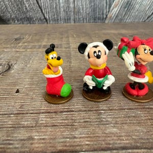 May include: Three small Disney character figurines. Pluto is in a red and green Christmas stocking. Mickey Mouse is wearing a red coat and a white hat. Minnie Mouse is wearing a red dress and holding a gift.