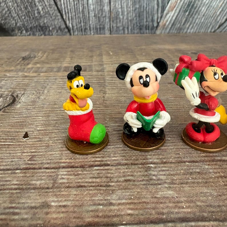 May include: Three small Disney character figurines. Pluto is in a red and green Christmas stocking. Mickey Mouse is wearing a red coat and a white hat. Minnie Mouse is wearing a red dress and holding a gift.
