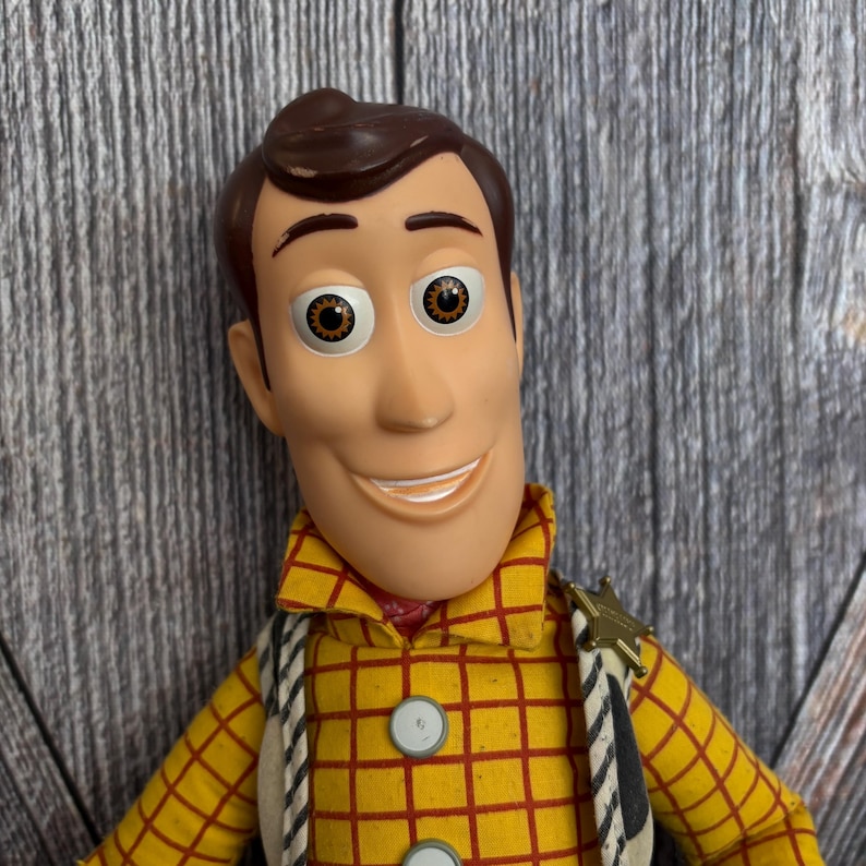 May include: A Woody action figure, with a smiling face, brown hair and eyes. He wears a yellow plaid shirt with red lines, a sheriff's badge, and a white vest. The figure is a collectible toy.