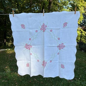 May include: White linen tablecloth with a scalloped edge, adorned with embroidered pink floral clusters and a central wreath design. Green stems connect the flowers. The tablecloth is hanging outside on a clothesline.