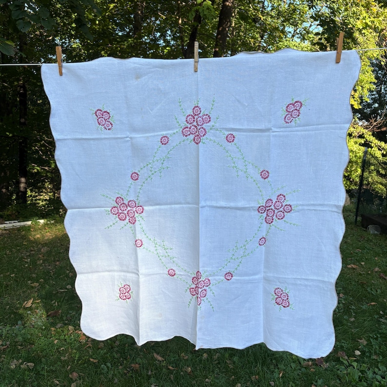 May include: White linen tablecloth with a scalloped edge, adorned with embroidered pink floral clusters and a central wreath design. Green stems connect the flowers. The tablecloth is hanging outside on a clothesline.