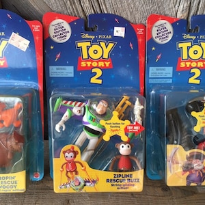 May include: Three Toy Story 2 action figures in their original packaging. The packaging is blue and red with the Toy Story 2 logo. One figure is Buzz Lightyear with a monkey, and the other two are Woody and a cowboy.
