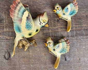May include: Three ceramic bluebirds with yellow and brown accents. The birds are painted with a detailed design and have a 3D effect. The birds are perched on a wooden surface.