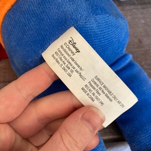 Puede incluir: Un juguete de peluche azul con una etiqueta blanca que dice "Disney" y "Manufactured under license by/ Fabriqu&eacute; sous licence par Just Play, LLC 4850 T-Rex Ave, Suite 100, Boca Raton, FL 33431, USA SURFACE WASHABLE ONLY. AIR DRY. Contents: All New Materials Polyester Fibers REG.NO. PA-15431 (HK) MADE IN CHINA"