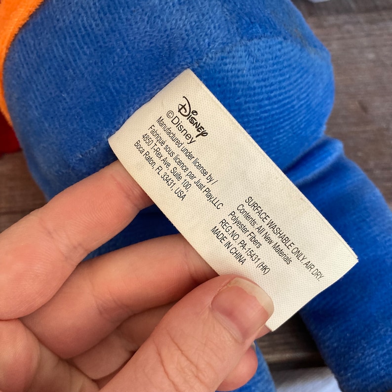 Puede incluir: Un juguete de peluche azul con una etiqueta blanca que dice "Disney" y "Manufactured under license by/ Fabriqu&eacute; sous licence par Just Play, LLC 4850 T-Rex Ave, Suite 100, Boca Raton, FL 33431, USA SURFACE WASHABLE ONLY. AIR DRY. Contents: All New Materials Polyester Fibers REG.NO. PA-15431 (HK) MADE IN CHINA"