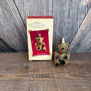 May include: A Hallmark Keepsake Threadbear ornament. The brown teddy bear wears a thimble hat and a green collar with a red heart. The ornament is next to its original box, which features the same bear design.