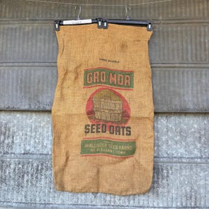 May include: A vintage burlap sack with the text "GRO-MOR SEED OATS" in green and red. The sack is suspended from a black hanger. The text "THREE BUSHELS" is above the logo, and "SOLD BY MCALLISTER SEED FARMS MT. PLEASANT, IOWA" is below.