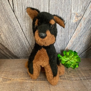 May include: A small black and brown plush dog toy. The dog is sitting with its paws tucked under its body. It has a black nose and brown eyes.