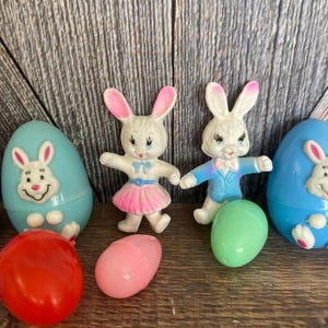 May include: A set of vintage Easter egg toys featuring bunny figurines and plastic eggs. The eggs are blue, pink, and green. The bunnies are white with pink and blue accents.