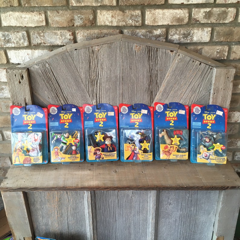 May include: Six Toy Story 2 action figure sets in their original packaging. Each package has a blue and red design with the Toy Story 2 logo. The figures are displayed on a wooden shelf against a brick wall.